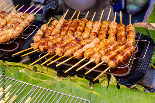 Foto Grilled fermented pork skewers cooking on a hot electric grill at a lively street food market, showcasing golden caramelized surfaces and authentic Thai cuisine