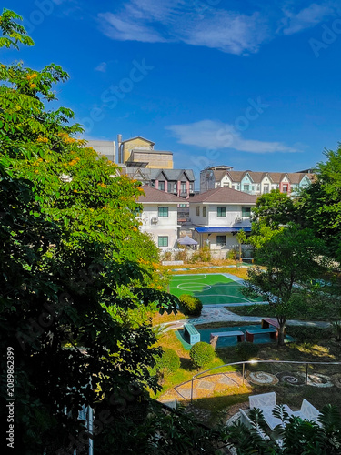 A vibrant outdoor view framed by lush green trees, featuring residential buildings under a bright blue sky. The peaceful urban landscape captures the beauty of nature blending seamlessly with modern a