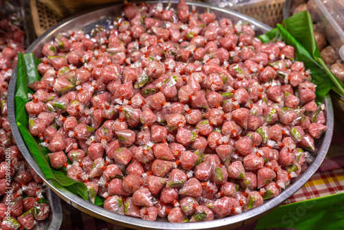 Traditional Thai fermented pork sausage individually wrapped in plastic and neatly arranged in a large stainless steel tray lined with banana leaves at a local market.