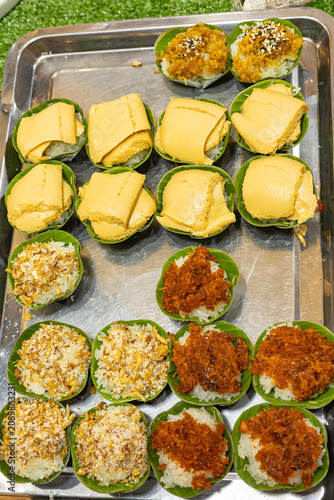 Assorted Thai sticky rice desserts arranged on a stainless steel tray, featuring pandan custard, shredded fish, and sweet shrimp toppings in banana leaf cups.