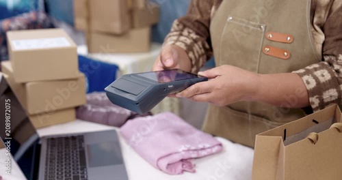 Elderly asian artisan online seller tilts POS terminal toward customer phone, payment approval icon appears, laptop business management runs behind, shibori textile parcels queued for delivery.