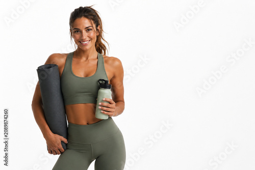 Fit and Fabulous: A smiling woman in athletic wear, holding a yoga mat and water bottle, embodies fitness and well-being, ready for her workout.