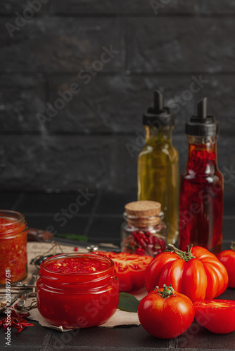 Homemade tomato sauces, ketchup and adjika in glass jars with heirloom tomatoes on dark background