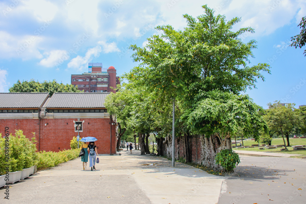 Obraz premium TAIPEI, TAIWAN – FEBRUARY 15, 2022: Tourists walking along a sunny pathway next to historical red brick buildings and green trees at Huashan 1914 Creative Park.