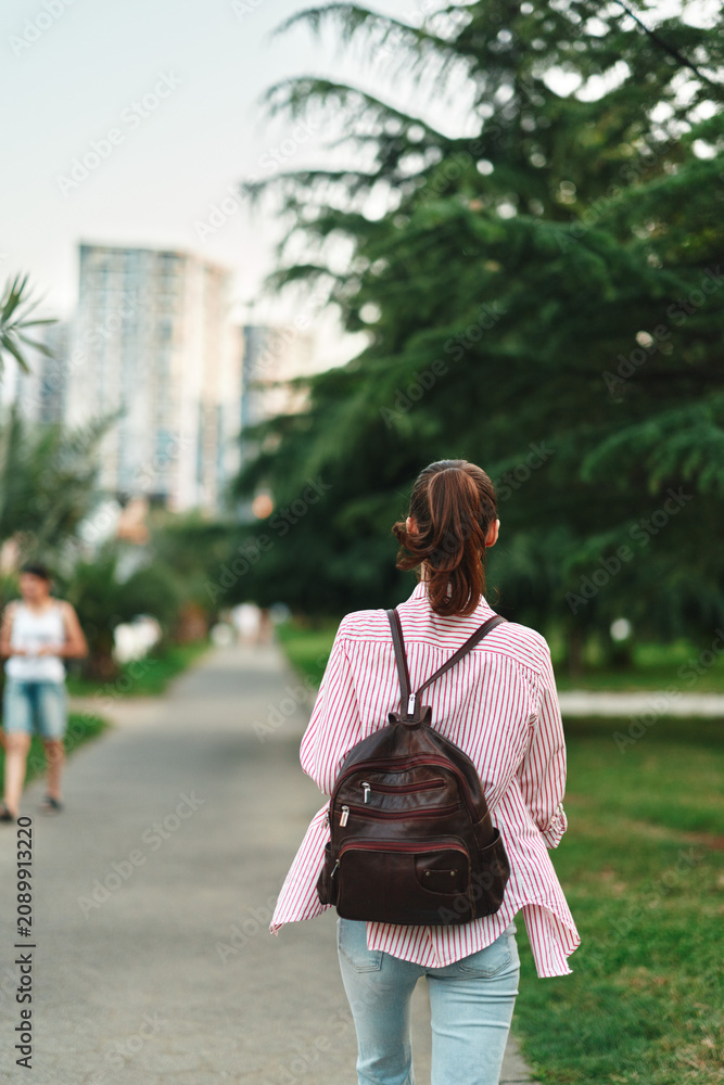 Naklejka premium Casual woman with backpack walks along a tree lined park path, city skyline in the distance, relaxed lifestyle mood, everyday outdoor exploration and gentle breeze on a sunny day