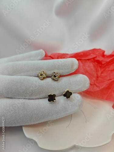Vertical photo of gloved hand holding two pairs of gold clover stud earrings. Plain gold and diamond set jewelry display