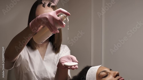 Professional beautician squeezing skincare toner or cleansing lotion on a cotton pad for a young african american woman in a beauty clinic. Facial treatment preparation