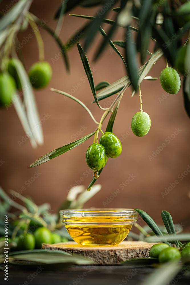 Naklejka premium Small glass bowl filled with olive oil placed on wooden surface surrounded by fresh green olives and olive branches. Concept of Mediterranean cuisine, organic farming, healthy food and natural harvest