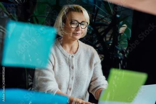 Stylish 50's confident mature businesswoman, middle aged company director, experienced senior female professional, business coach working on laptop in office. View through glass wall with sticky notes