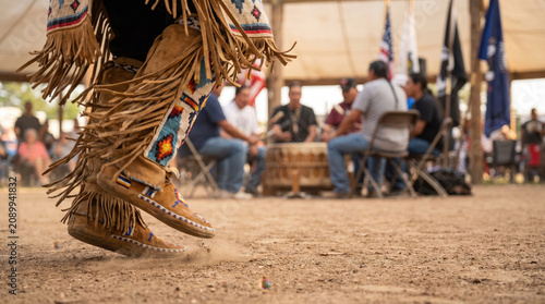 Native dance regalia moccasin fringe pow wow footstep raise dust during cultural ceremony with drum circle and community gathering under open shelter