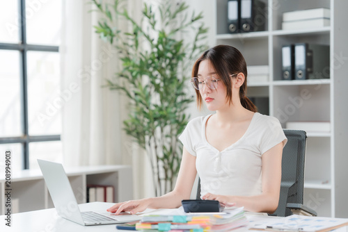 Valokuva Young Woman Working at Desk: A focused young woman with glasses sits at her desk, typing on a laptop and surrounded by office supplies, suggesting a productive workday or study session