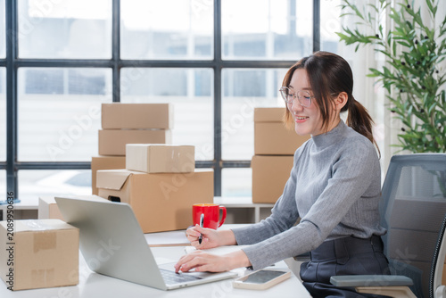 Bild auf Leinwand E-commerce Entrepreneur at Work: A young woman, an aspiring entrepreneur, diligently works at her laptop surrounded by shipping boxes, suggesting a small business operation
