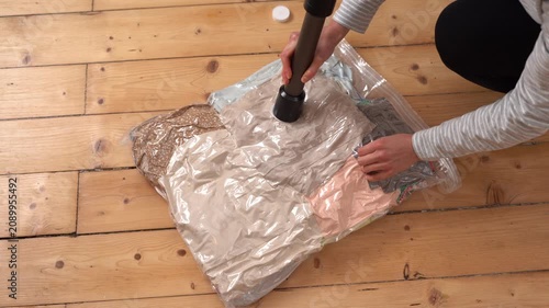 A woman uses a vacuum cleaner to remove air from a space saver bag full of clothes. The bag compresses, shrinking the garments for compact storage or travel packing. Filmed on a wooden floor