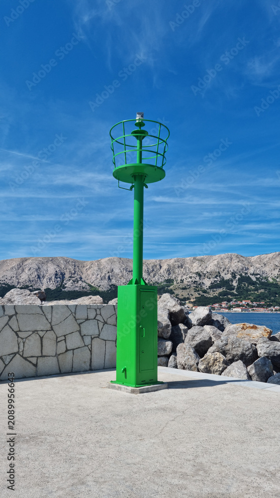 Fototapeta premium A vibrant green navigation lighthouse tower standing on a concrete pier in Baska on Krk island with mountains in the background.