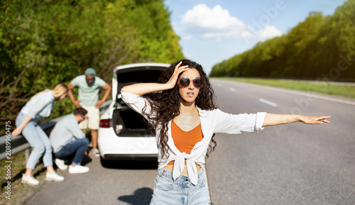 Принти на полотні Millennial lady trying to stop passing car, asking for help, her friends fixing broken vehicle, having road accident