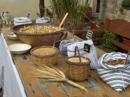 A rustic table setting of Cretan fotokolyva salad ingredients with Greek labels, featuring grains, legumes, baskets, wheat stalks, and a white cat in the background.