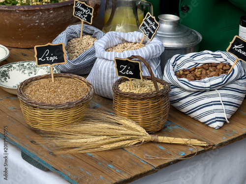 A rustic display of Cretan fotokolyva ingredients in baskets and sacks with Greek labels for wheat, zea, broad beans, manarolia, and black-eyed peas, next to wheat stalks.
