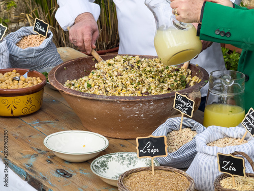 Pouring fresh lemon-oil dressing over traditional Cretan fotokolyva, a healthy Mediterranean vegan salad. Bowls of raw ingredients with Greek labels like 'Sitari' (wheat) surround the table.