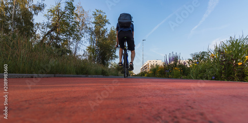 Konstfotografi Low angle back view of a man with a backpack riding a bicycle on a red asphalt b