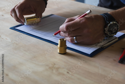 High angle shot of a worker writing with a red pencil on a clipboard chart next to brass components on a wooden table. Quality control