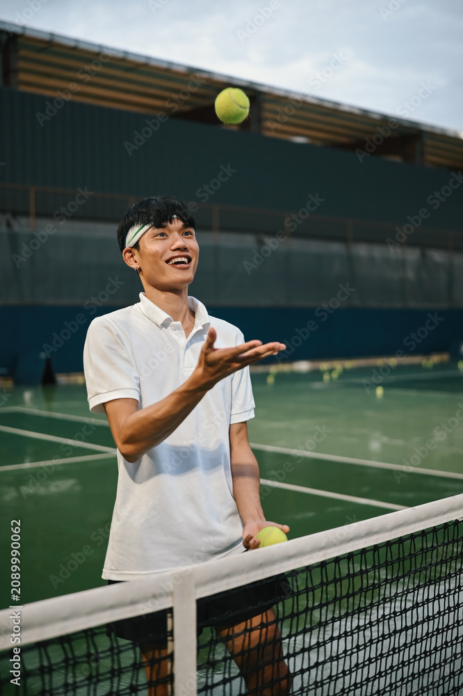 Obraz premium Young Asian Man Smiling and Tossing Tennis Ball at Net on Outdoor Court