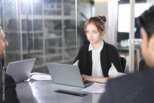 Tableau sur toile Businesswoman with a bun glances aside from her laptop as a dark-haired male colleague leans in from the foreground during a discussion in a bright glass-walled coworking office