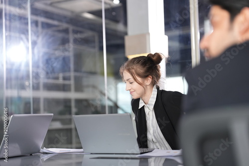 Obraz na plátně Businesswoman with a bun smiles gently while typing on her laptop as a dark-haired colleague leans in from the foreground in a modern glass-walled coworking office in Bangkok