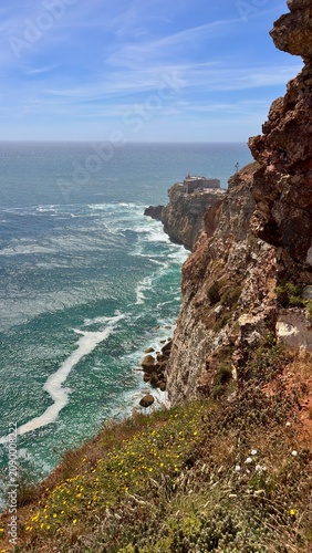 View of the ocean waves crashing against the cliffs near a coastal town under a blue sky and bright sunlight, Portugal