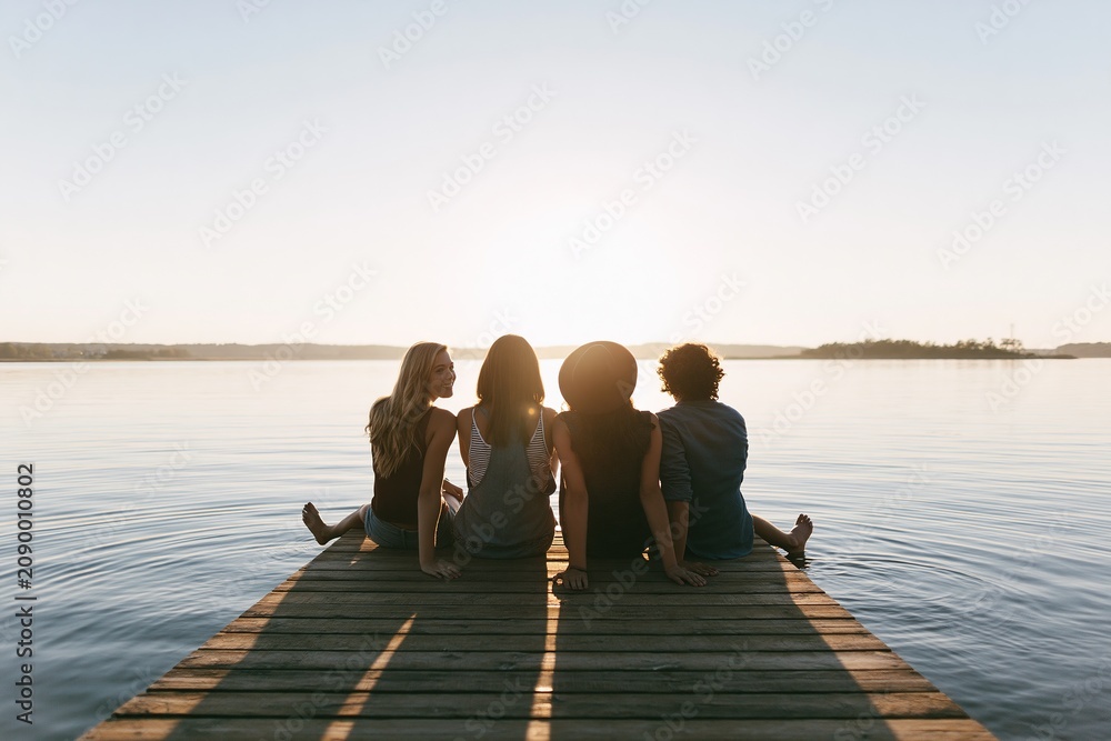 Fototapeta premium Four Friends Relaxing on Dock by Lake at Sunset