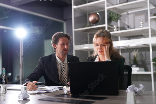 Fotografie Businessman points at a document explaining his fix while the woman holds her head and listens at the paper strewn night desk in a bright modern coworking office