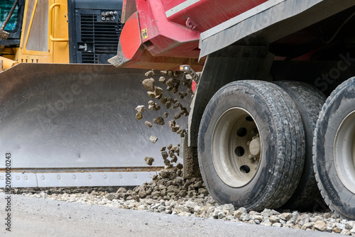Close-up of a heavy dump truck unloading crushed stone or gravel in front of a bulldozer metal blade during road construction.