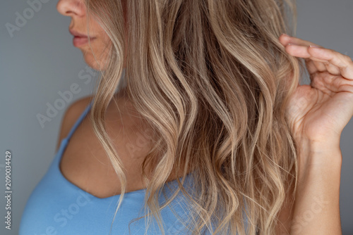 Close-up of wavy blonde hair with soft highlights, young woman touching healthy textured hair after styling or coloring on a grey background
