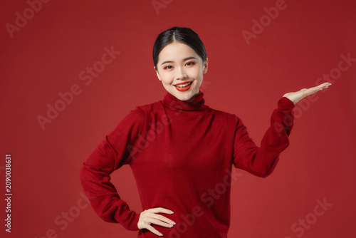Photography Smiling young woman in red sweater presenting with open palm on red background