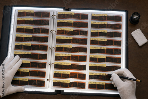 Overhead view of gloved hands labeling organized film negatives in archival binder sleeves on a light table with lens and cleaning cloth.