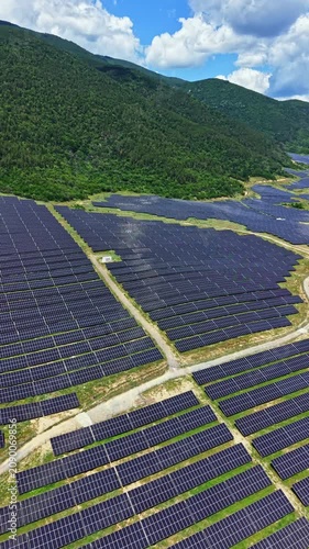 Rows of solar panels stretch across a wide area under a blue sky with fluffy clouds. The green hills surround the field, showing efforts to harness renewable energy.