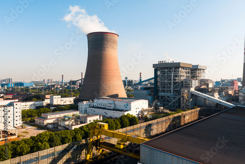 Schilderij op canvas Aerial view of a chinese nuclear power plant cooling tower emitting steam near a modern city, featuring surrounding industrial railways and solar panels at sunrise