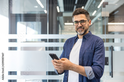 Confident mature businessman wearing glasses and a denim shirt, smiling at the camera while using a smartphone in a modern corporate office environment