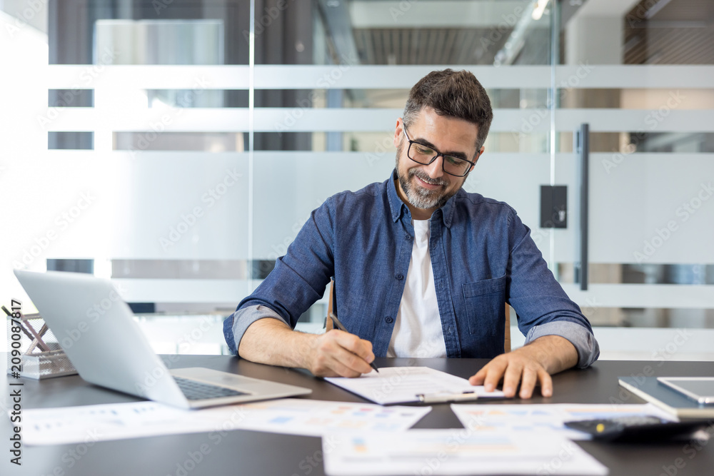 Obraz premium Concentrated businessman in casual attire reading and signing an important contract or document, sitting at a modern office desk with a laptop and paperwork