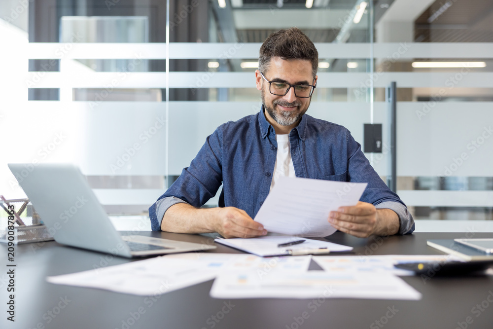 Fototapeta premium Smiling businessman reviewing paperwork and financial reports at his office desk, symbolizing careful planning, corporate success, and meticulous business management