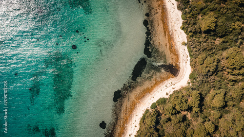 Curved rocky shoreline beside turquoise sea and dense green forest seen from high drone view on sunny coastal day with sparkling water and visible shallow stones near narrow pebble beach