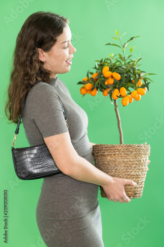 Fotografía Young woman stands against green screen, holding woven pot with green kumquat pl