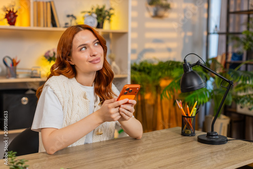 Young woman on home table typing smartphone messages during remote work with focused calm mood and steady nods. Adult girl on home table chatting and working from home in cozy living room work mode.