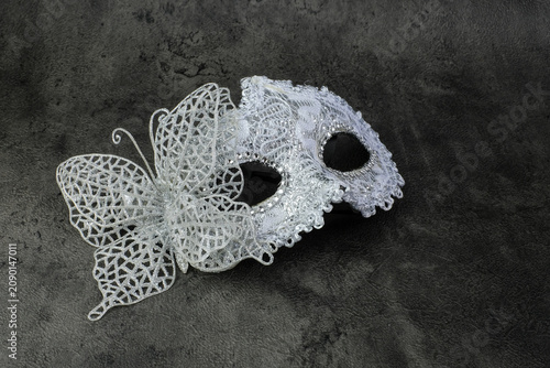 A silver lace masquerade mask with a large butterfly ornament rests on a dark grey textured background. Suitable for masquerade party, carnival costume, Halloween decoration, mystery theme