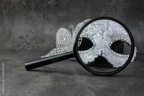 A magnifying glass lies in front of a silver lace masquerade mask on a dark textured background. Suitable for hidden identity, mystery concept, detective theme, masquerade party