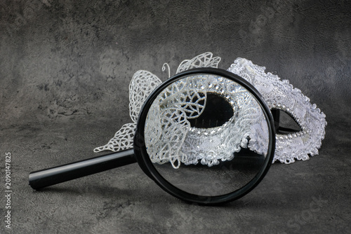 A black magnifying glass frames part of a silver butterfly masquerade mask on a dark grey surface. Suitable for mystery identity, masquerade investigation, hidden face