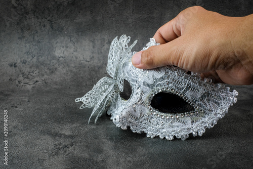 A hand holds a silver lace masquerade mask with butterfly decoration on a dark grey textured background. Suitable for masquerade preparation, carnival costume, Halloween party