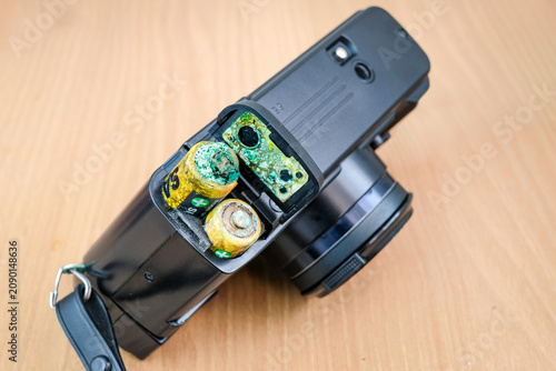 Corroded batteries are visible inside an open battery compartment of a black electronic device on a wooden table. Suitable for corroded flashlight batteries, battery leak damage, electronic failure