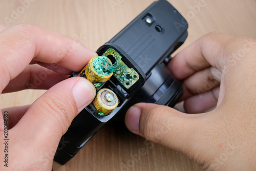 Hands remove corroded batteries from an open electronic device on a wooden table. Suitable for battery replacement, leaking battery repair, electronic maintenance, corrosion cleanup