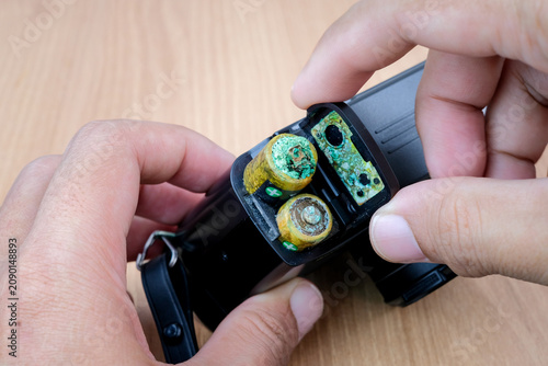 Hands open a battery compartment with corroded leaking batteries inside an electronic device. Suitable for battery leakage, damaged electronics, repair warning, corrosion problem