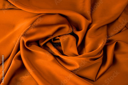 Orange satin fabric forms a soft swirl pattern with smooth folds and highlights. Suitable for luxury textile background, festive design, fashion fabric, warm backdrop, or product display content.